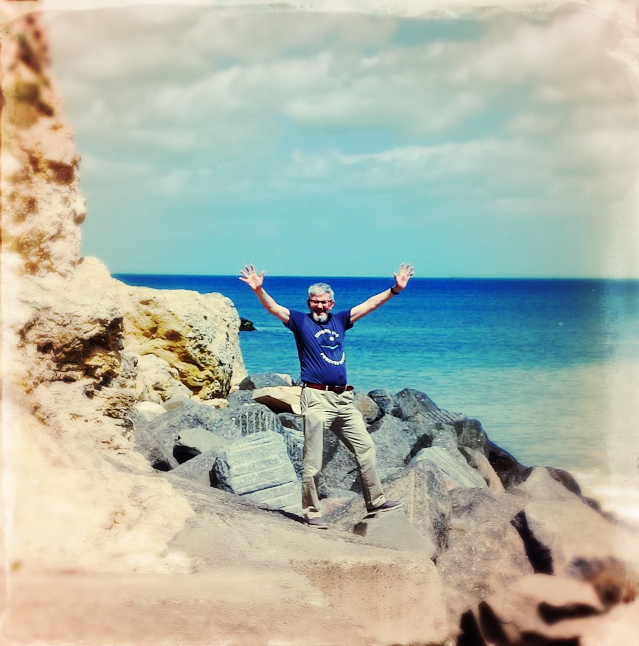 older man, on rocks near the sea, with arms upraised in happiness